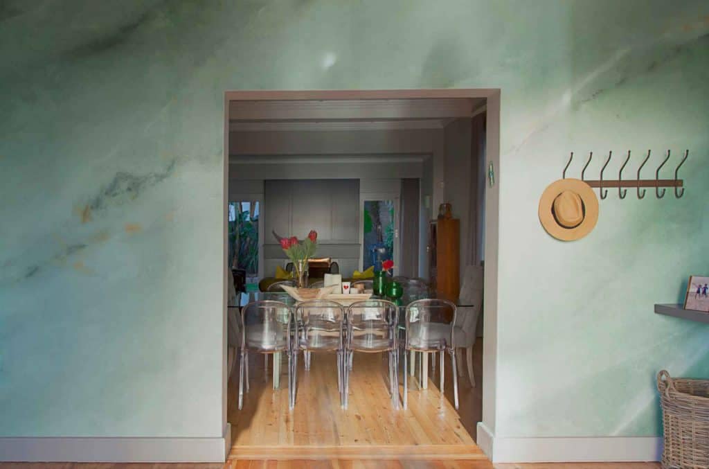 The wallpaper features a luxurious green marble pattern with intricate white veining, adding sophistication to the modern dining room. Hints of gold accents seamlessly blend into the design, providing a touch of elegance. This striking backdrop enhances the overall aesthetic by complementing clear plastic chairs and a wooden dining table perfectly. The subtle interplay of colors and textures in this wallpaper makes it ideal for high-end interior design showcases in an online portfolio.