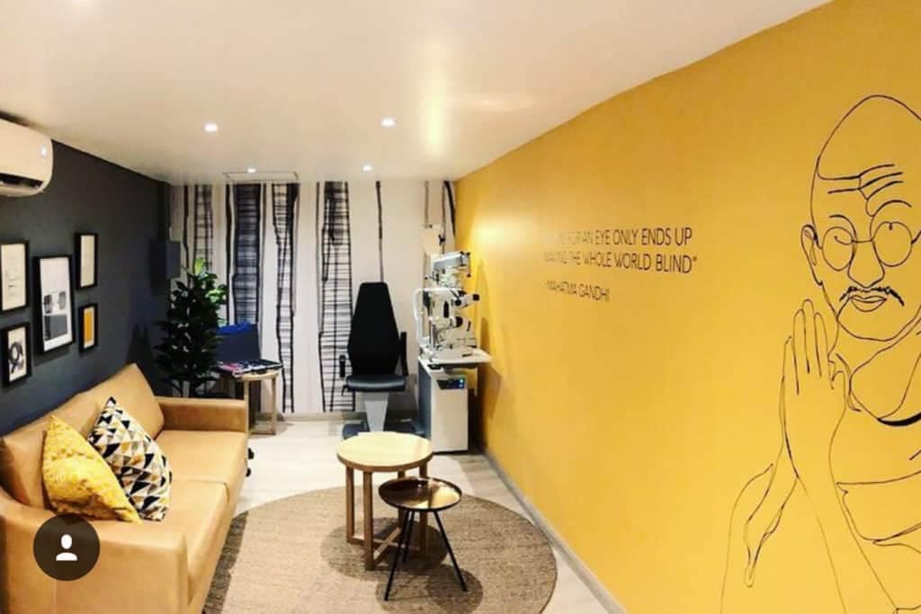 A modern eye clinic waiting room features a vibrant yellow wall adorned with a line drawing of Mahatma Gandhi. The wallpaper highlights elements like the intricate sketch and the inspiring quote, adding warmth and positivity to the space. The bright yellow background creates an inviting atmosphere, ideal for a contemporary yet comforting setting.