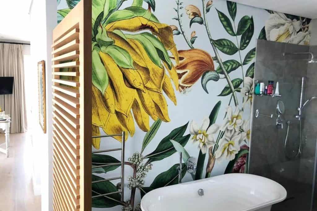 Tropical botanical-themed wall mural with lush green palm leaves, vibrant monstera plants, colorful hibiscus flowers in shades of pink and red, and exotic birds like toucans and parrots. Rich greens, lively pinks, deep reds, and accents of yellow dominate the design.