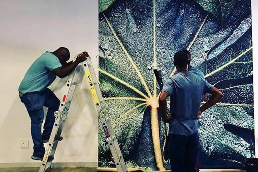 A large, detailed image of a water-covered leaf, showcasing vibrant green hues with glistening water droplets, is being hung on the wall. The wallpaper features intricate textures and shades that capture the delicate veins of the leaf and the reflective quality of the water.