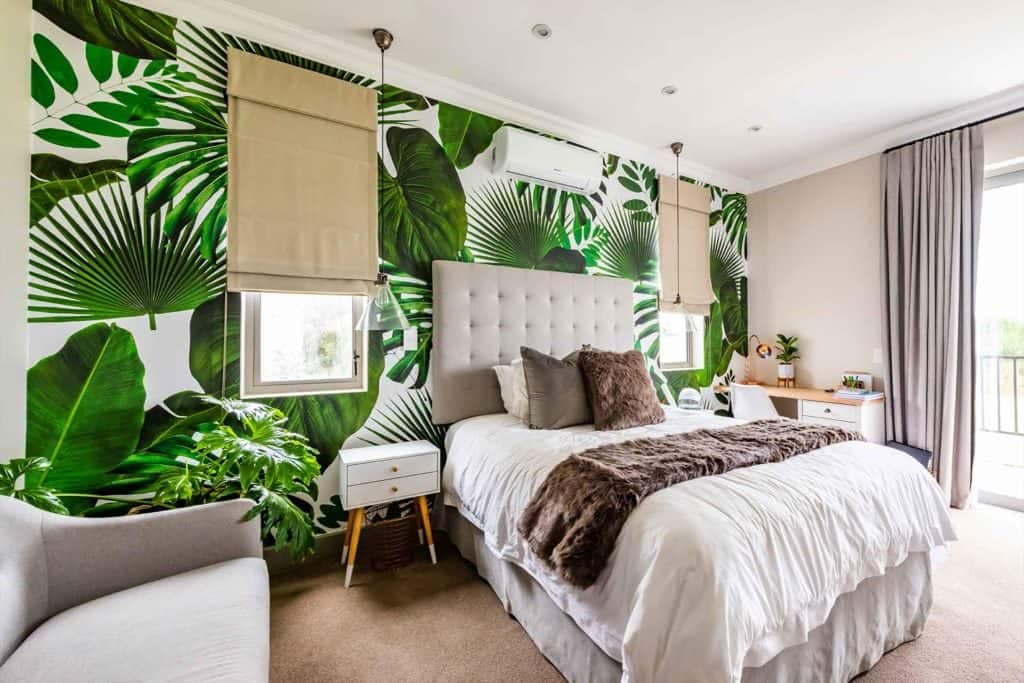 The wallpaper features a tropical theme with large, vibrant green leaves that create an immersive, lush atmosphere. The pattern includes various shades of green and subtle accents of earthy tones to mimic natural foliage. The design is bold yet elegant, providing a striking backdrop for the bedroom's decor.