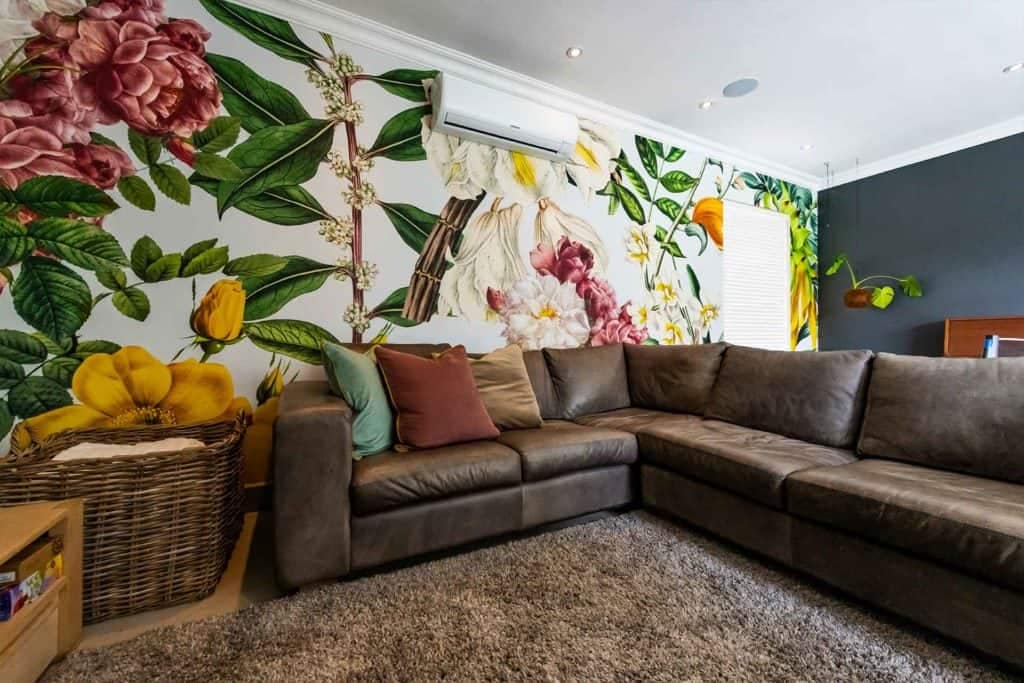 The wallpaper features a vibrant floral mural showcasing various flowers in shades of pink, yellow, and blue with green leaves. The intricate design includes blossoms and foliage against a lively background. Perfect for adding a burst of color and nature-inspired beauty to any space, this floral wallpaper is both eye-catching and elegant.