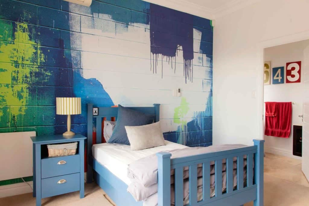 The wallpaper in the bedroom features a dynamic blue and green abstract mural. The design is bold with rich hues of blue and vibrant shades of green, blending seamlessly to create an artistic backdrop. This visually striking pattern sets a lively tone for the room.