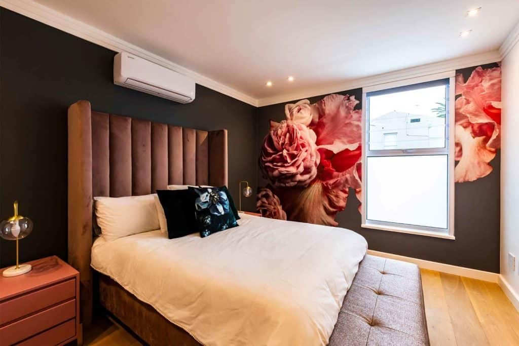 The modern bedroom's wallpaper features a large, intricate floral mural in rich, vibrant colors against a dark background. Highlighted elements include oversized blossoms in shades of pink, white, and deep green foliage. The dramatic contrast and detailed botanical design make it a striking focal point.