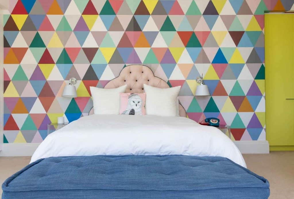 This bedroom features a vibrant geometric triangle-patterned wallpaper. The design includes bold triangles in shades of blue, pink, yellow, green, and white. Each triangle is distinct yet harmonizes with the entire pattern, creating an eye-catching and dynamic backdrop for the room's decor.