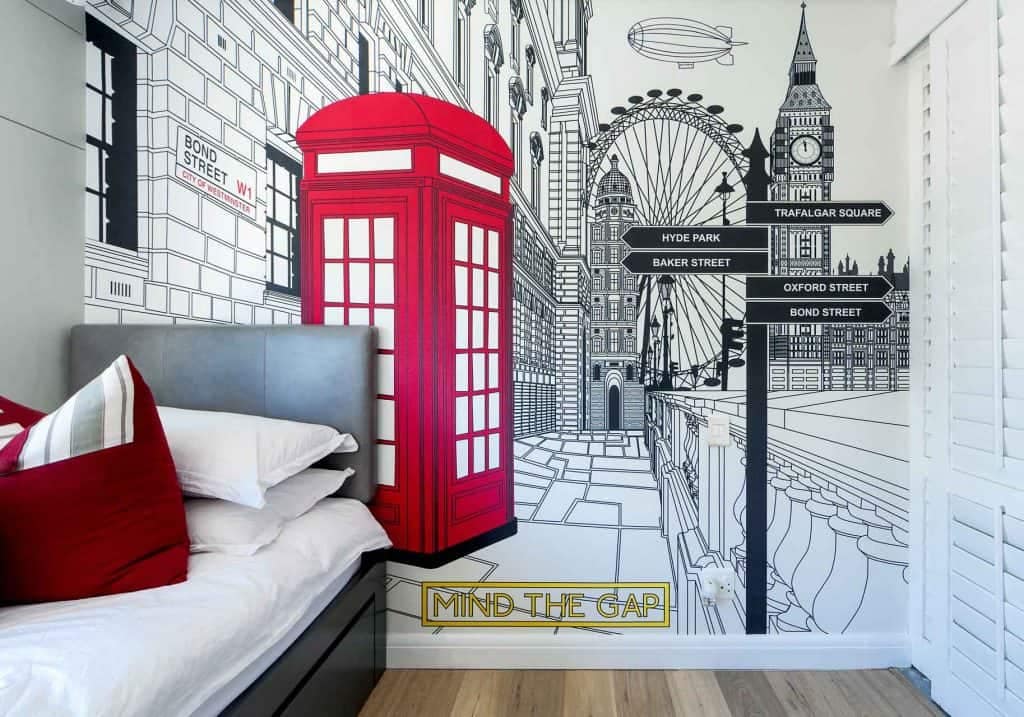 The bedroom wallpaper is a detailed mural featuring iconic London landmarks such as Big Ben with its recognizable clock face, the London Eye's giant wheel, and a classic red telephone booth. The background includes a collage of street signs from various famous locations around London. On the floor near the bed, the phrase "Mind the Gap" is prominently displayed in bold letters. The mural uses vibrant colors: rich reds for the telephone booth, deep blues for the street signs, golden tones on Big Ben, and shades of white and gray for the cityscape elements.