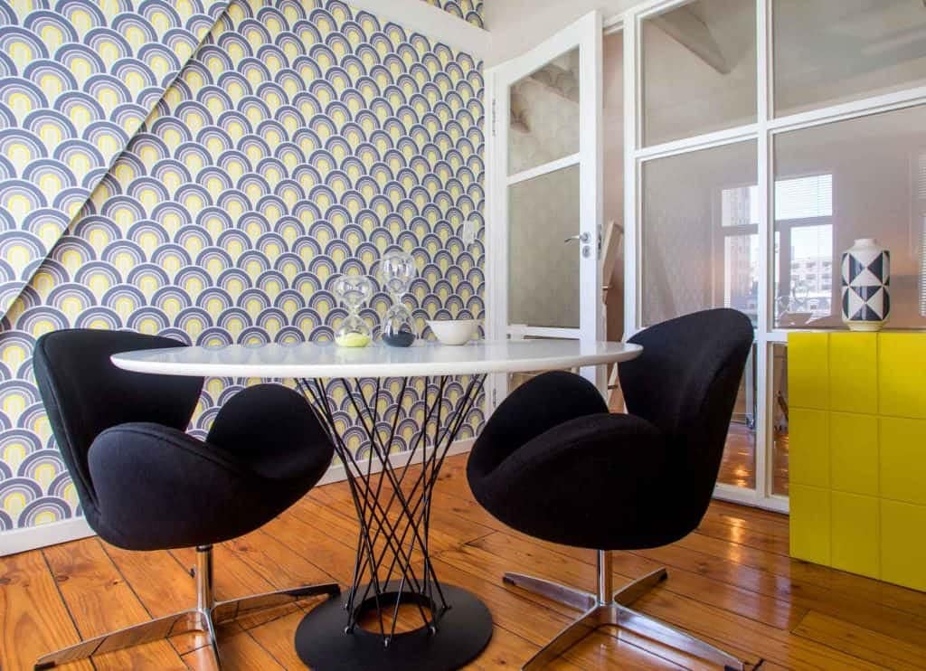 The wallpaper in this modern dining area features a vibrant yellow and blue patterned design. It incorporates geometric shapes and intricate motifs, creating a lively and eye-catching backdrop. The interplay of bold yellows with various shades of blue adds depth and visual interest, making it an ideal choice for enhancing the space's aesthetics.