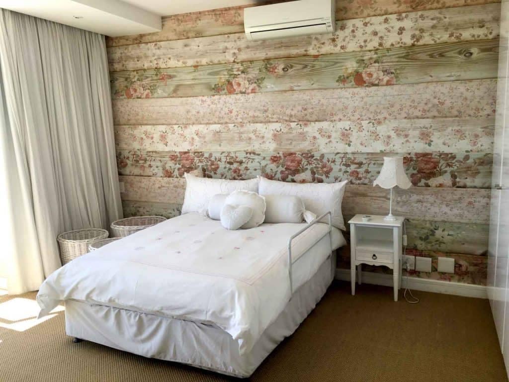 The floral-patterned wallpaper is the focal point, featuring a blend of soft pastels and bright hues. The design includes delicate pink roses, lush green leaves, and subtle hints of lavender. This charming pattern adds warmth and texture to the wooden wall, creating a cozy yet vibrant atmosphere perfect for a bedroom interior.