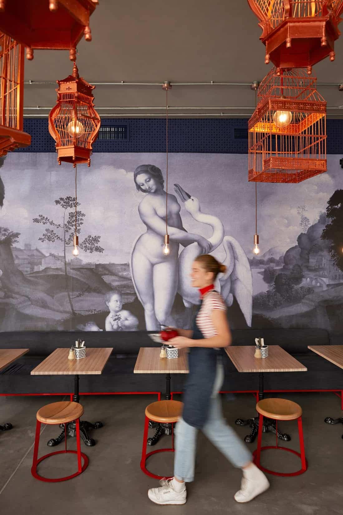 A dimly lit restaurant, featuring a large mural on the wall depicting a nude figure and a swan. The mural's colors include earthy tones, soft beiges for the figure, and white with subtle gradients for the swan. Red birdcage-shaped pendant lights are notable ceiling decor, casting a romantic glow. This artistic wallpaper combines classical elements with modern touches, suitable for an elegant yet contemporary ambiance.