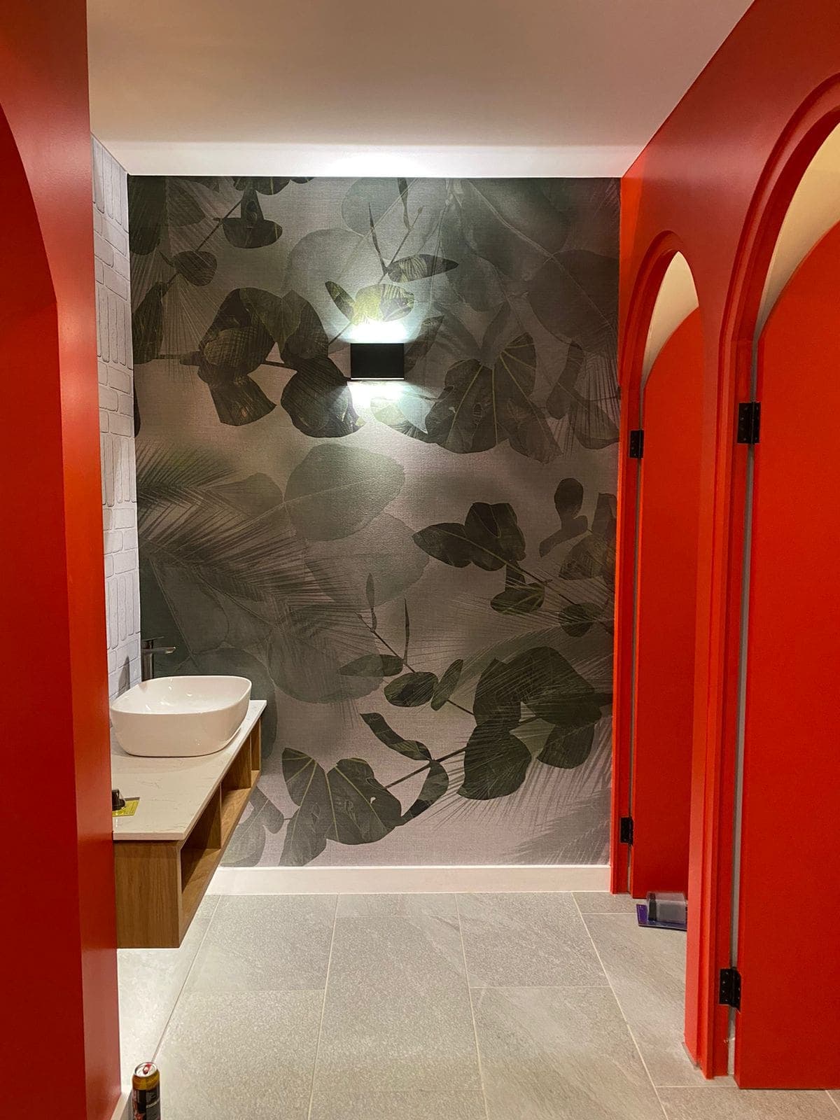 The vibrant bathroom features a back wall adorned with a large leafy wallpaper, showcasing rich green foliage with intricate vein patterns. Hints of earthy browns and subtle yellows accentuate the natural design, creating a lush, verdant backdrop. This lively botanical print adds dimension and visual interest to the space.