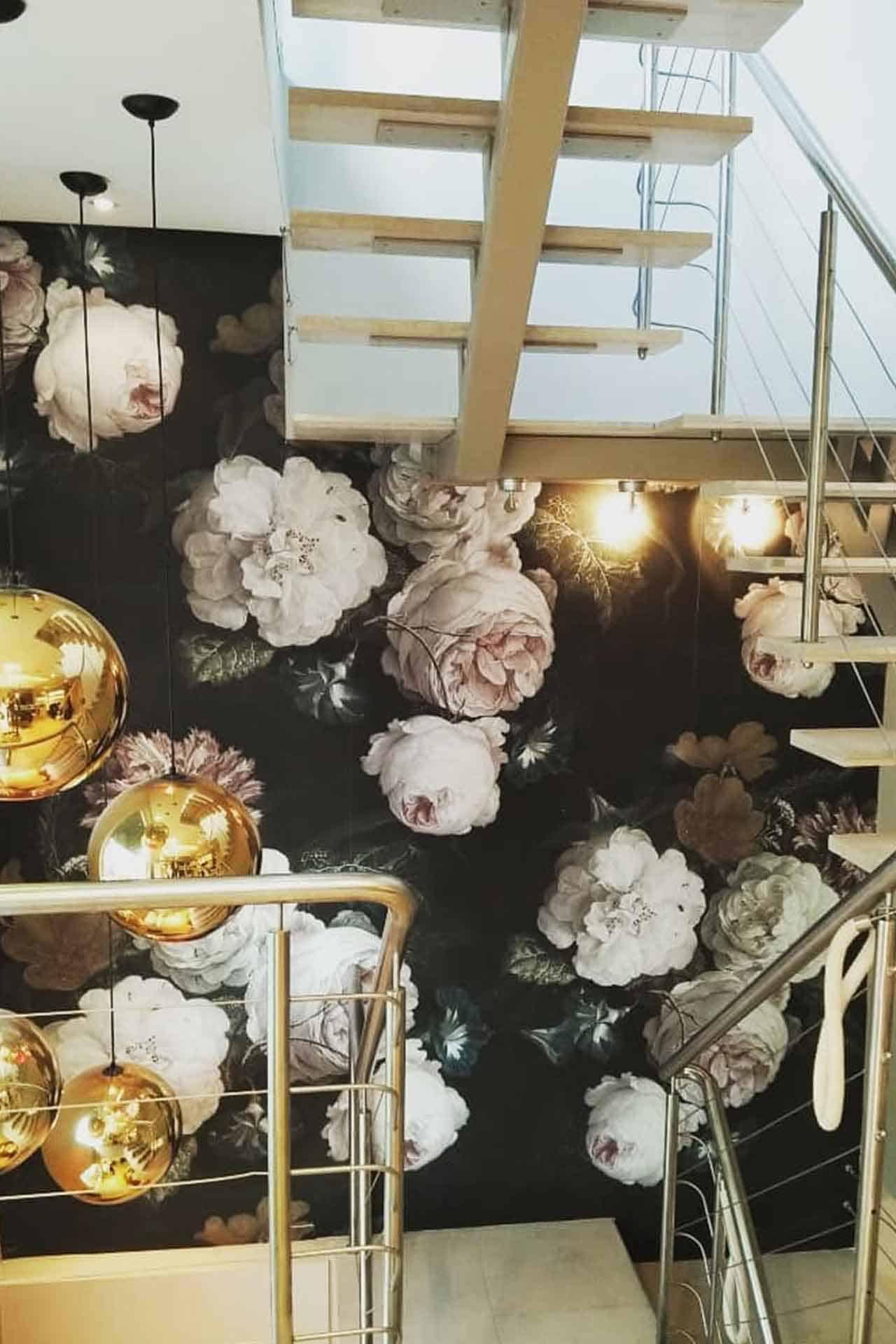 A large, dark floral-patterned wallpaper featuring white and pink flowers. The contrasted backdrop elevates the delicate hues of the blossoms, highlighted by illumination from three gold spherical pendant lights. This dramatic yet elegant design makes it an eye-catching feature for any portfolio.