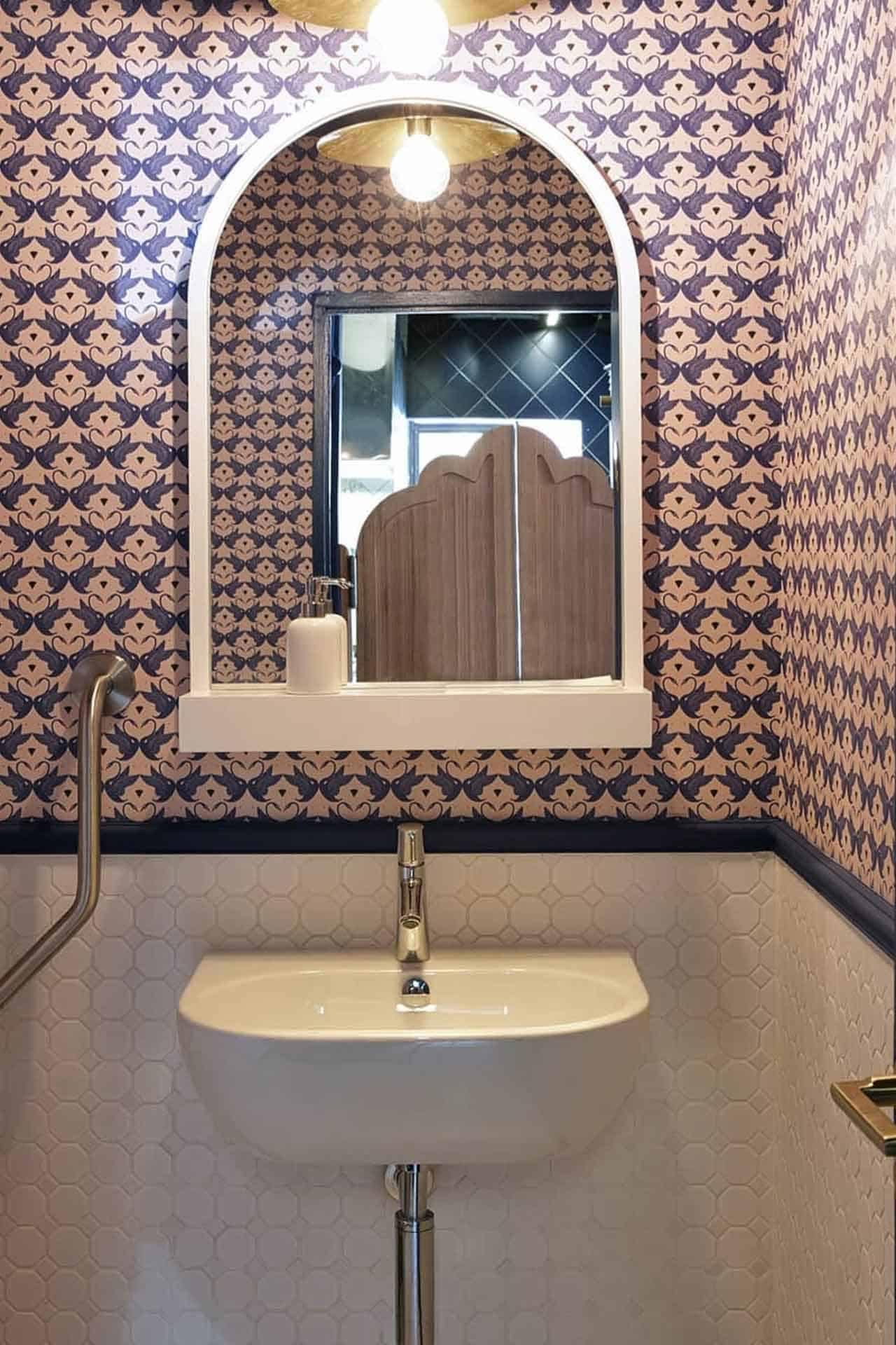 The wallpaper in the small bathroom is patterned with intricate floral designs featuring soft pastel shades of blue, pink, and green. Accents of gold add a touch of elegance to the pattern. The detailed botanical motifs create a visually striking backdrop in this cozy space.