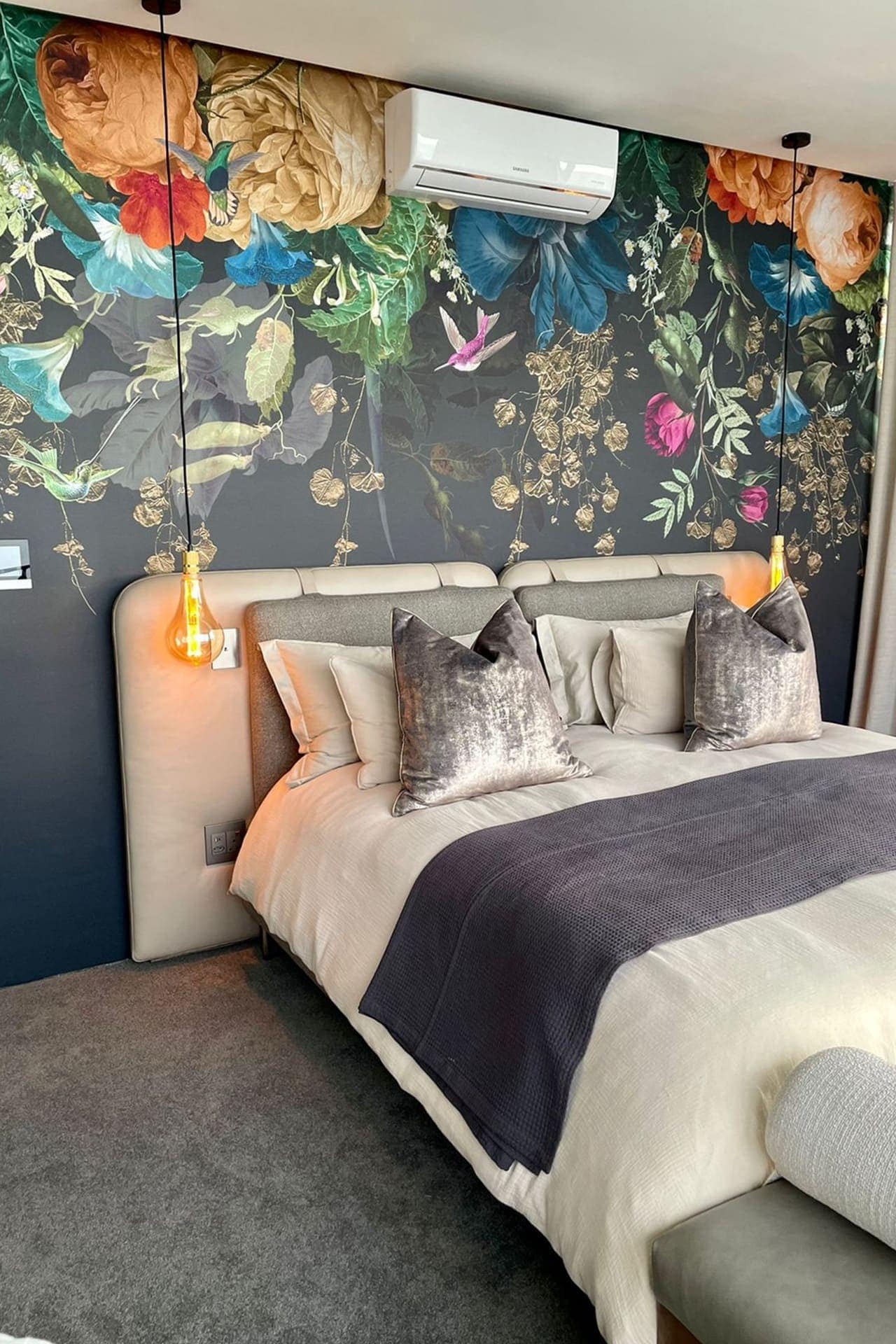 The wallpaper features a large floral mural in vibrant colors, including rich reds, deep purples, bright yellows, lush greens, and subtle blues. The intricate design showcases a variety of flowers and leaves, creating a bold and lively backdrop for the bedroom.