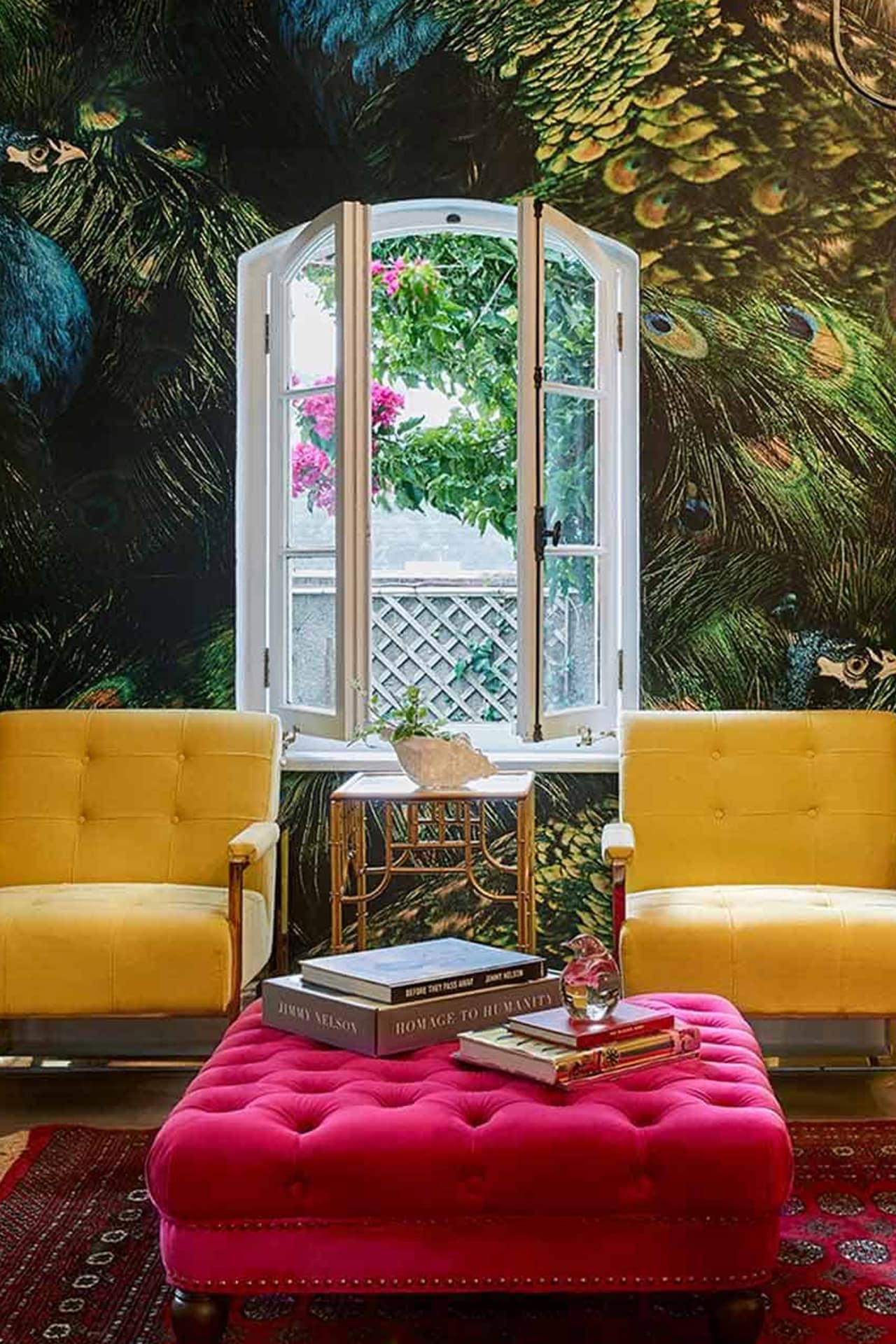 The wallpaper showcases a large, colorful peacock feather design, featuring vibrant hues of deep blues, rich greens, striking golds, and hints of iridescent purples. The intricate patterns highlight the feather’s eye-catching "eye" with concentric circles of turquoise, royal blue, and emerald. Each feather is detailed with fine lines and shimmering highlights that add depth and texture to the overall aesthetic. The lively mix of colors enhances the room's energy and complements the bright decor elements perfectly.