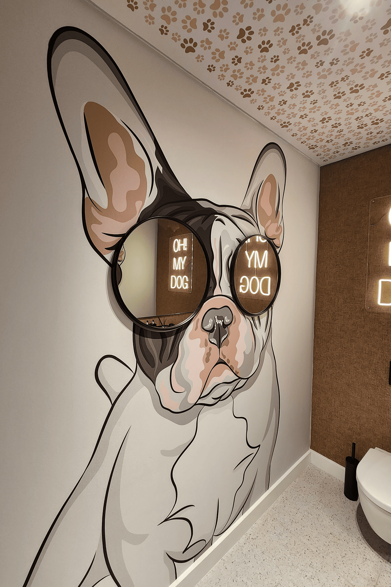 The wallpaper features a painted mural of a French Bulldog wearing large, reflective sunglasses with the text "OH MY DOG." The design includes a ceiling adorned with paw print patterns. Colors include various shades of gray for the bulldog, metallic hues for the sunglasses, and white for the text. The paw prints on the ceiling are black against a lighter background.