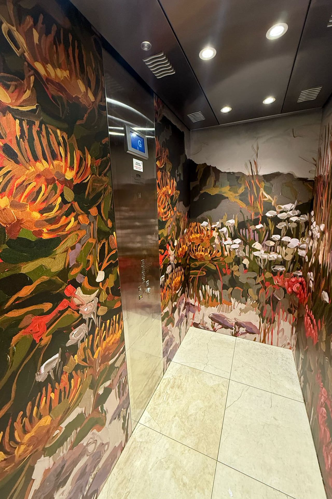 The elevator's interior is wrapped in a vibrant floral and plant-themed wallpaper. The design showcases bold, colorful abstract depictions of various flowers like lilies, roses, and daisies intertwined with lush greenery. Shades of deep magenta, rich emerald green, sunny yellow, soft lavender, and vibrant coral dominate the pattern. Intricate vines and leaves weave through the scene with accents in bright turquoise and gentle creams to add contrast to the lively floral motifs. This striking display creates an enchanting garden ambiance within the confined space.