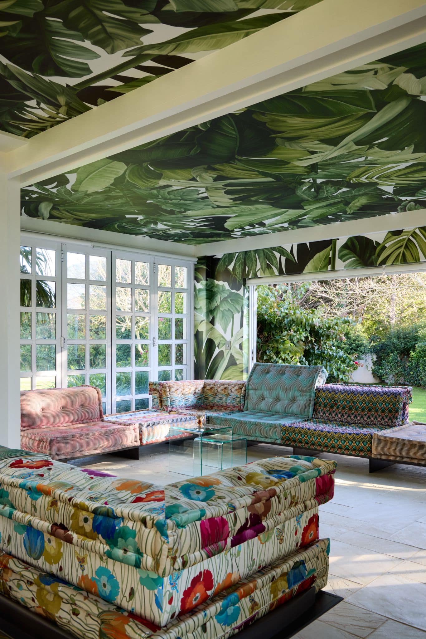 The sunroom features bold tropical leaf-print wallpaper covering the ceiling and walls, showcasing large green palm, banana, and monstera leaves with vibrant shades of green on a light background.
