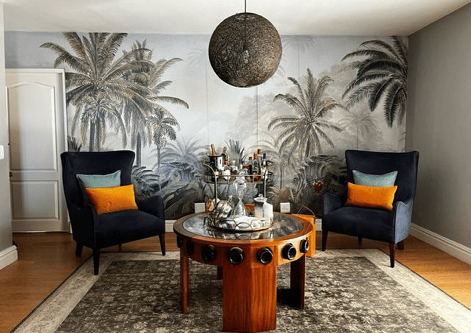 A stylish living room showcases a tropical-themed wallpaper featuring vibrant green palm trees, lush foliage, and exotic patterns. The backdrop incorporates shades of emerald and lime green with touches of earthy browns, evoking an island paradise feel.