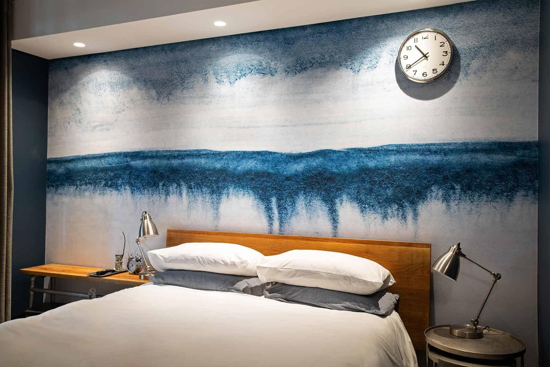 A modern bedroom with a stunning blue abstract wall mural behind the bed. The wallpaper features dynamic swirls and splashes of blue hues, accented by hints of white and subtle metallic touches. Perfect for portfolios, the mural adds a vibrant yet sophisticated statement to the space.