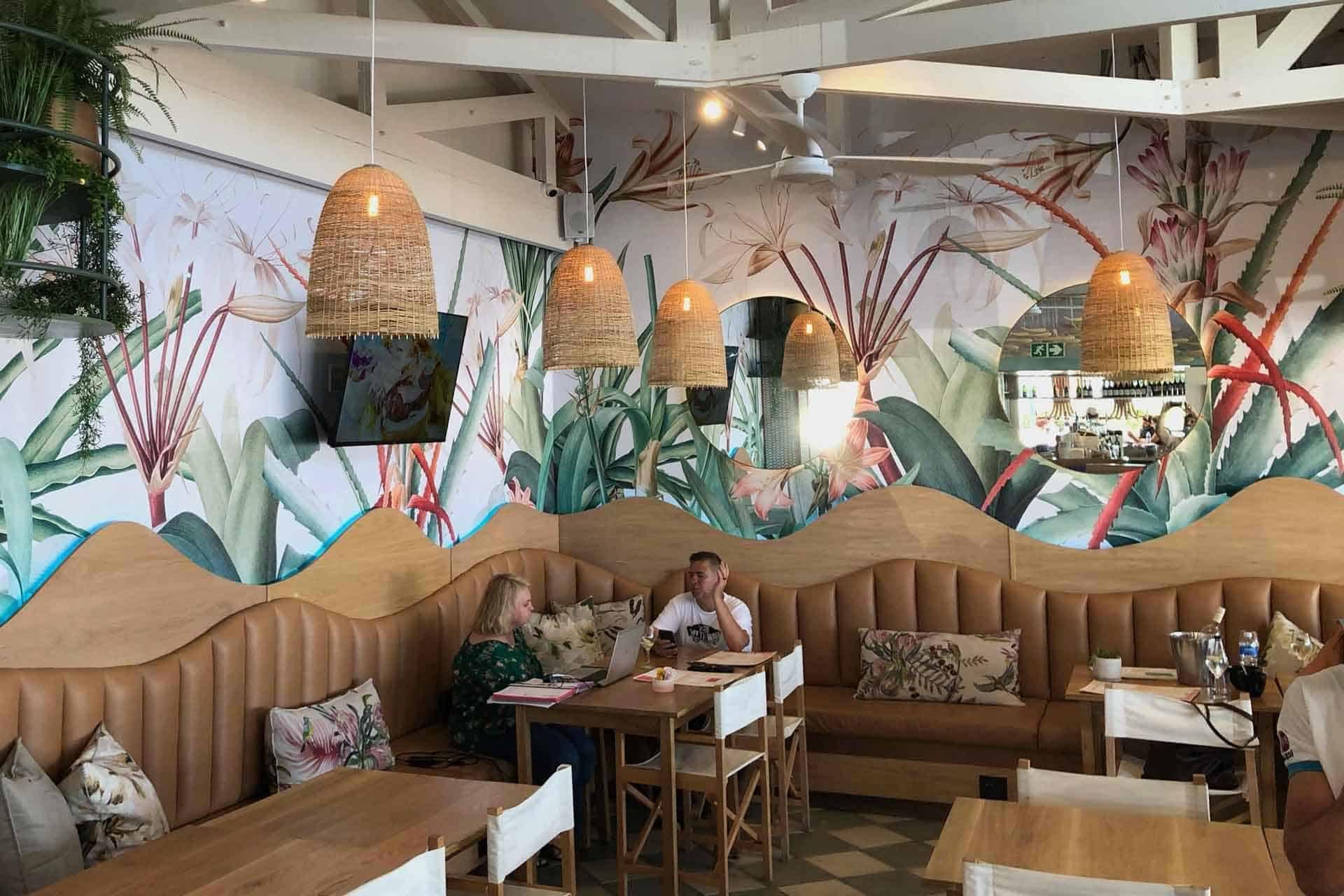 The wallpaper features vibrant tropical murals with lush greenery, including large palm leaves and colorful birds. The dominant colors are shades of green, bright blues, yellows, and splashes of red. There are also tropical flowers in pinks and oranges interspersed throughout the design.