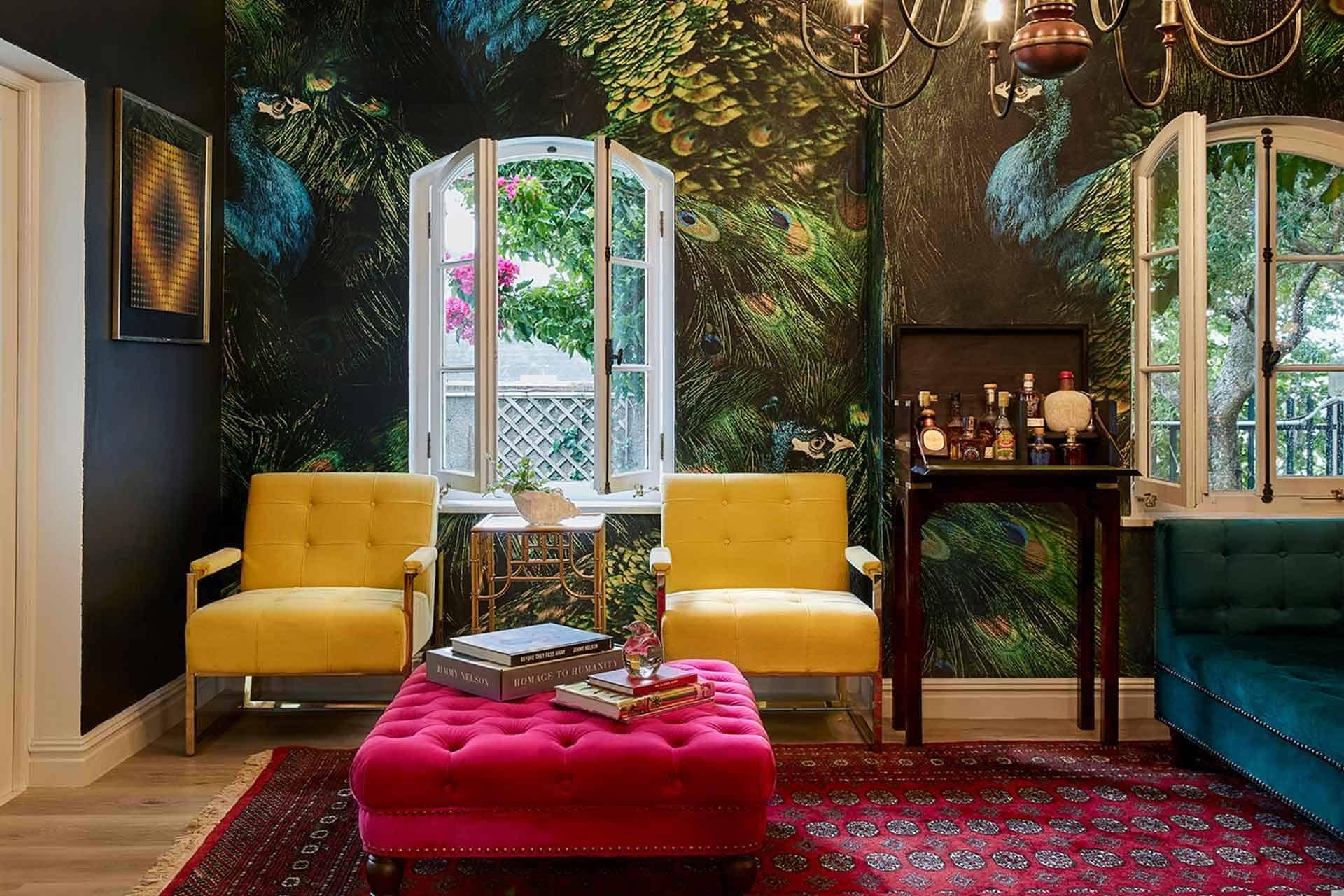 The wallpaper features an intricate peacock mural with vibrant hues of blue, green, and gold. It showcases detailed feathers with accents of iridescent teal, sapphire, emerald, and hints of deep indigo. The design includes lush greenery and ornate flourishes in rich shades of jade and chartreuse. Perfect for making a vivid statement wall in any living room décor portfolio.