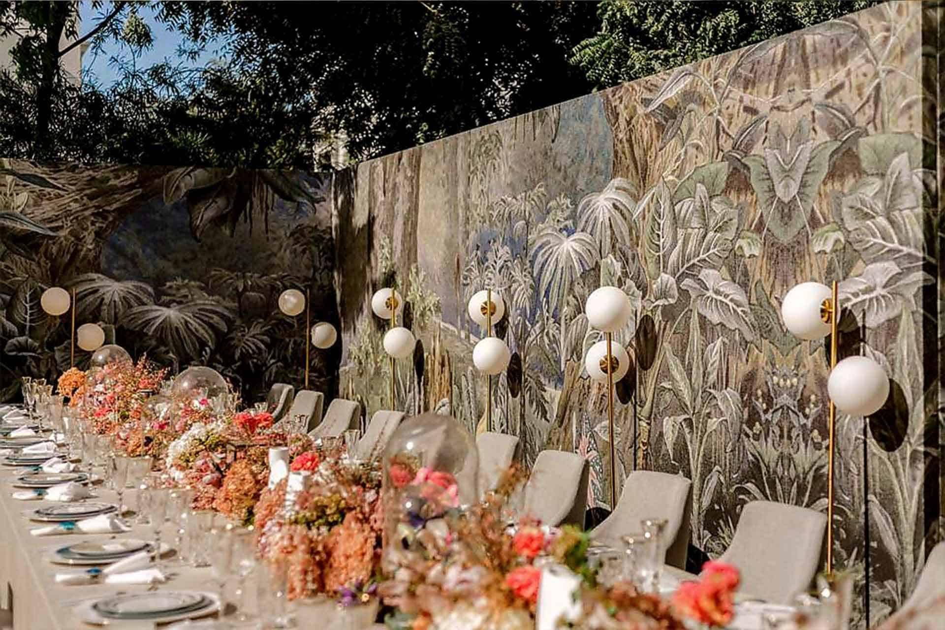 The wallpaper features a detailed mural with lush greenery and various plants. Shades of green dominate, complemented by earthy tones of brown and touches of vibrant colors from flowers. The intricate designs include leaves, vines, and botanical elements that enhance the natural theme.