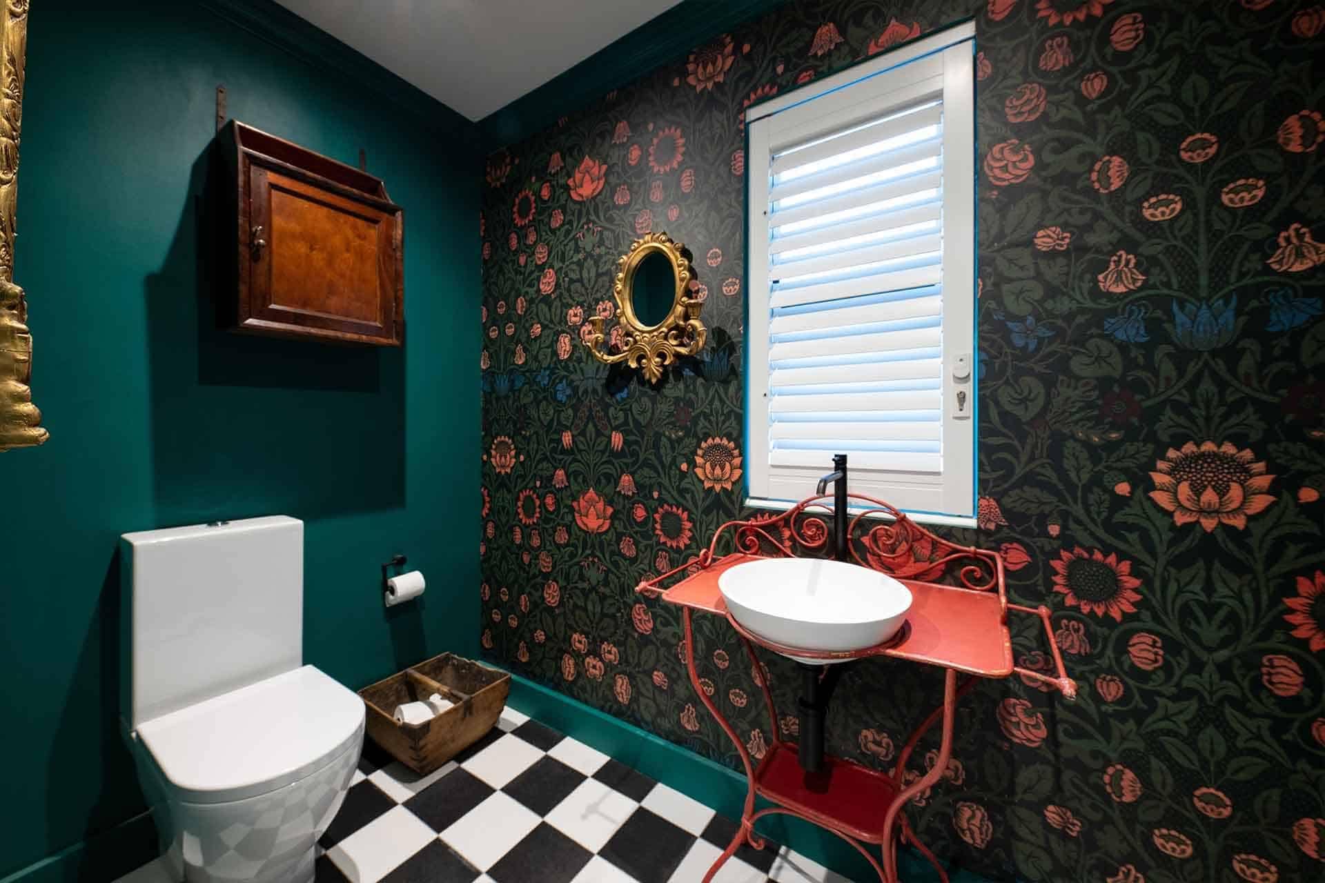 Dark floral wallpaper adorned with rich hues of deep reds, lush greens, and delicate pinks. It features intricate patterns of blooming flowers intertwined with leafy vines, creating a striking and elegant backdrop in the bathroom.