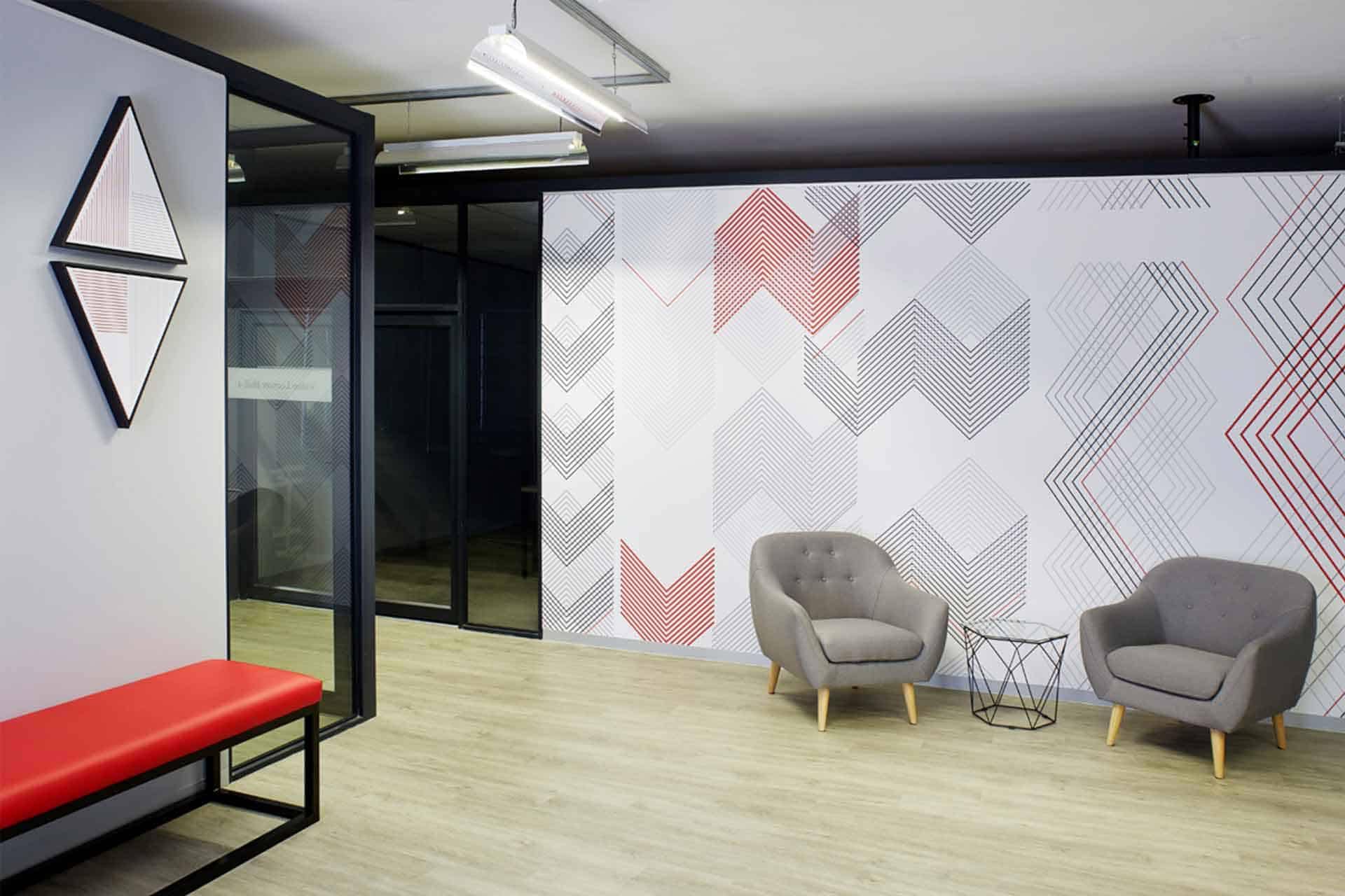 Modern office lounge features geometric-patterned wallpaper in red and gray tones. The design includes intricate shapes and lines, merging vivid red hues with subtle grays, creating a dynamic backdrop. Keywords: geometric-patterned wallpaper, red and gray tones, intricate shapes and lines.