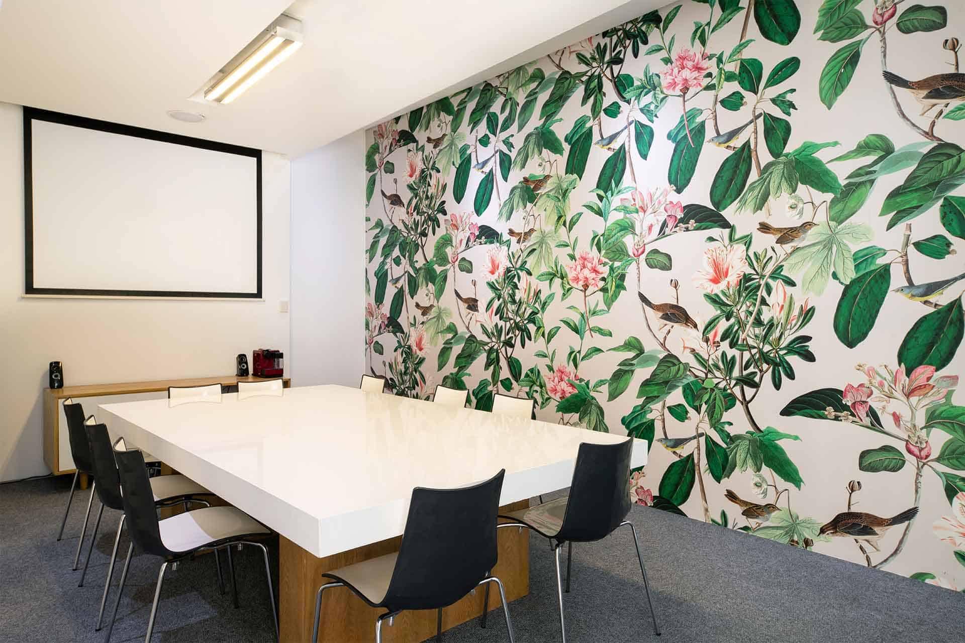 The wallpaper showcases a tropical-themed mural bursting with vibrant hues. Dominant elements include luxuriant green leaves and striking pink flowers, creating a lush, exotic vibe. This radiant combination of colors and shapes evokes an energizing jungle feel in the conference room.