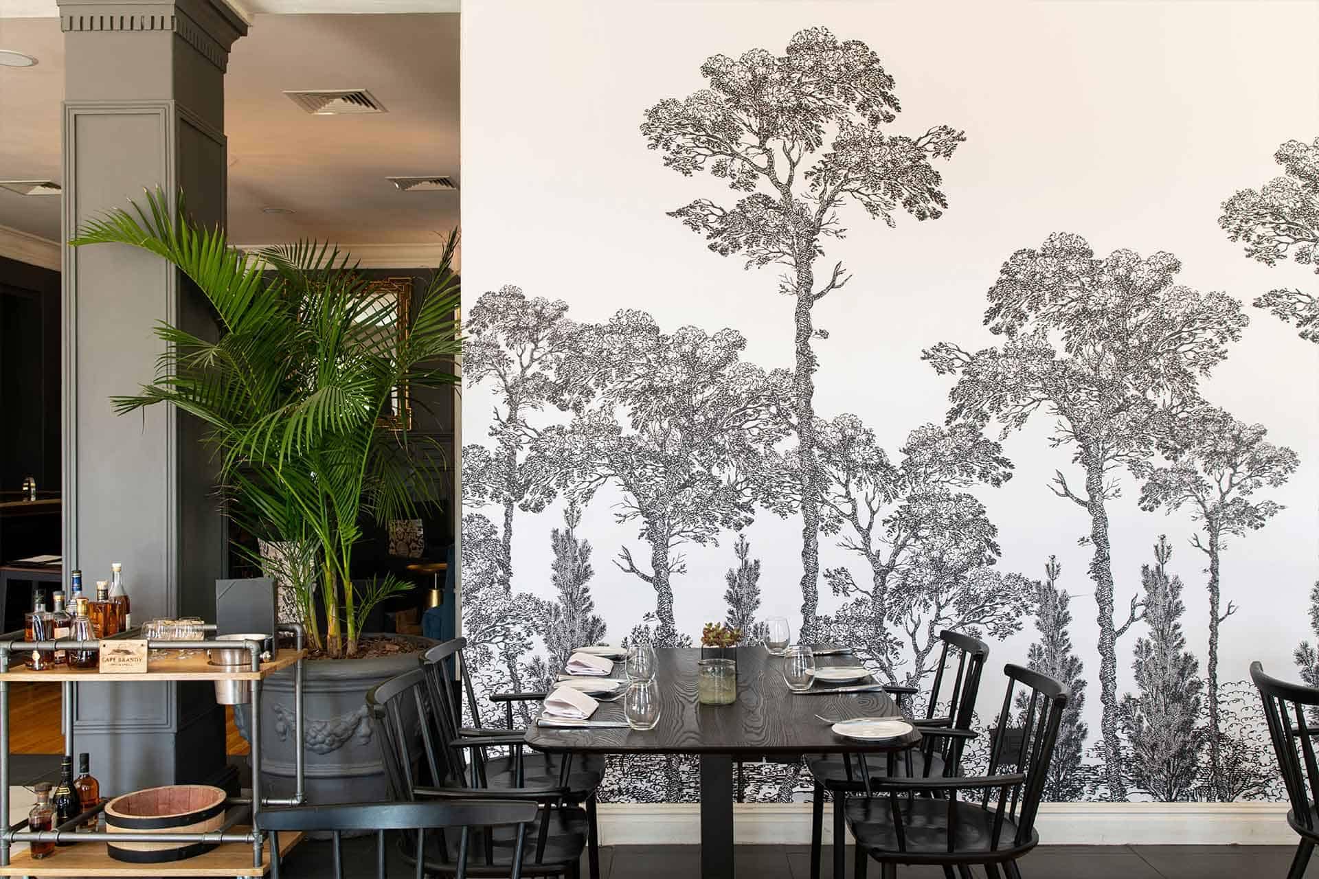 Modern dining area featuring a monochrome forest mural. The wallpaper showcases intricate details of trees and foliage in shades of black, white, and grey. The elegant design adds depth and a contemporary touch to the space, making it a striking focal point for any interior setting.