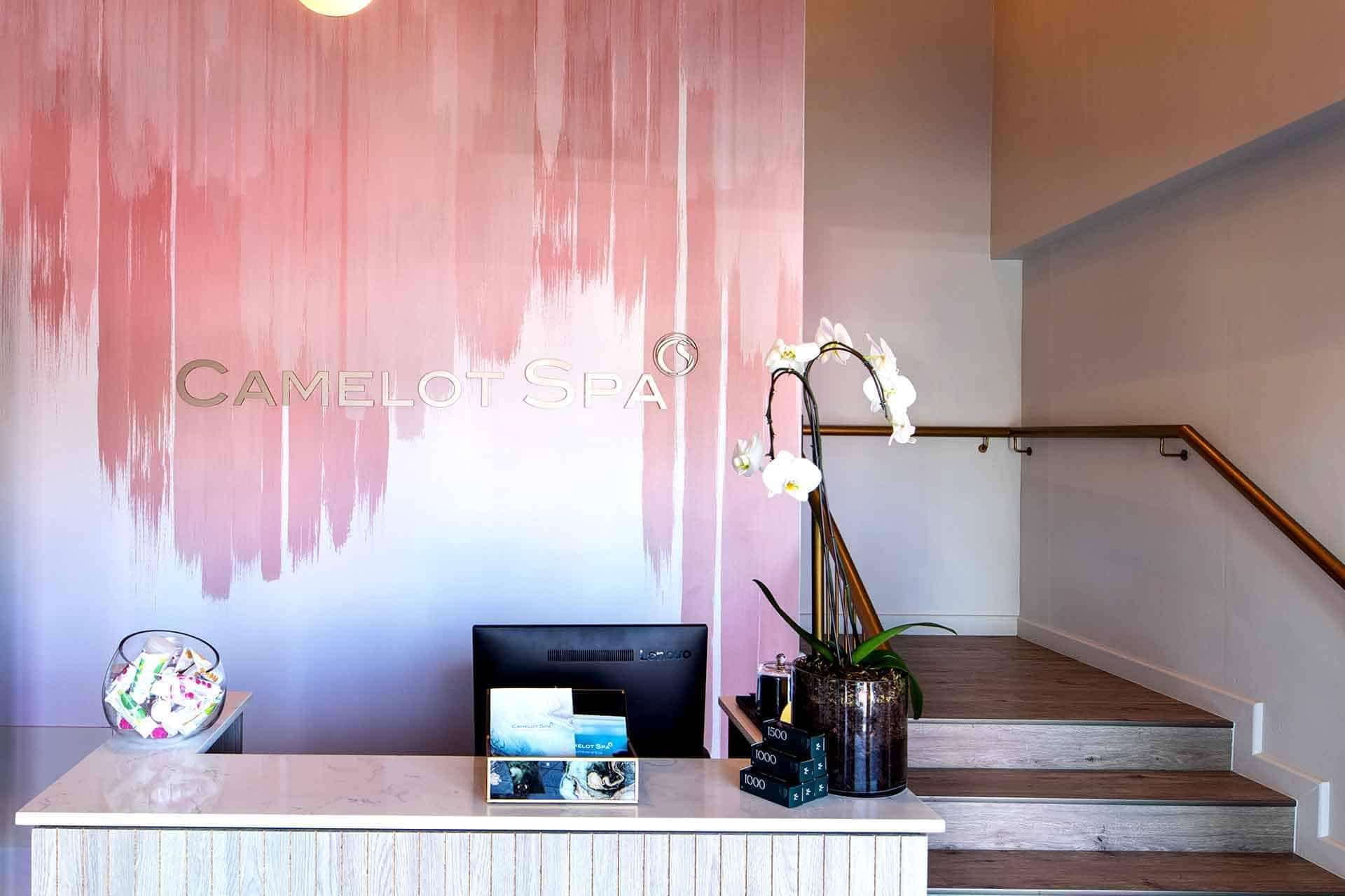 Modern gradient wall behind the front desk at Camelot Spa displays the spa's name. The wallpaper features a sleek transition from deep teal at the bottom to a soothing light blue at the top. The design includes subtle, elegant patterns resembling abstract, flowing waves and soft, delicate curves in various shades of blue and green.