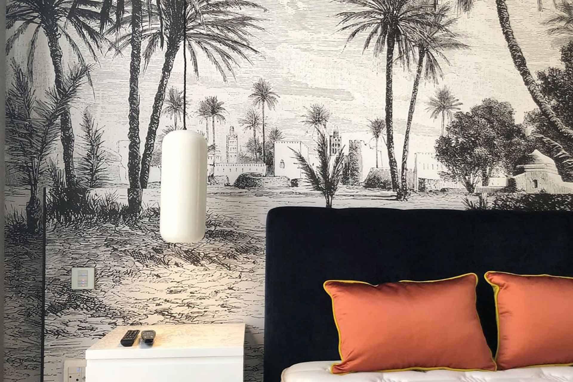 A bedroom featuring an intricate black and white wall mural of a desert scene with palm trees and buildings. The detailed wallpaper showcases elegant monochrome elements, including palm fronds, architectural structures, sandy dunes, and cloud-filled skies. This stunning visual piece creates a stark yet soothing backdrop that exudes artistic sophistication. Perfect for personal websites or portfolios seeking to highlight classic decor themes with contemporary flair.