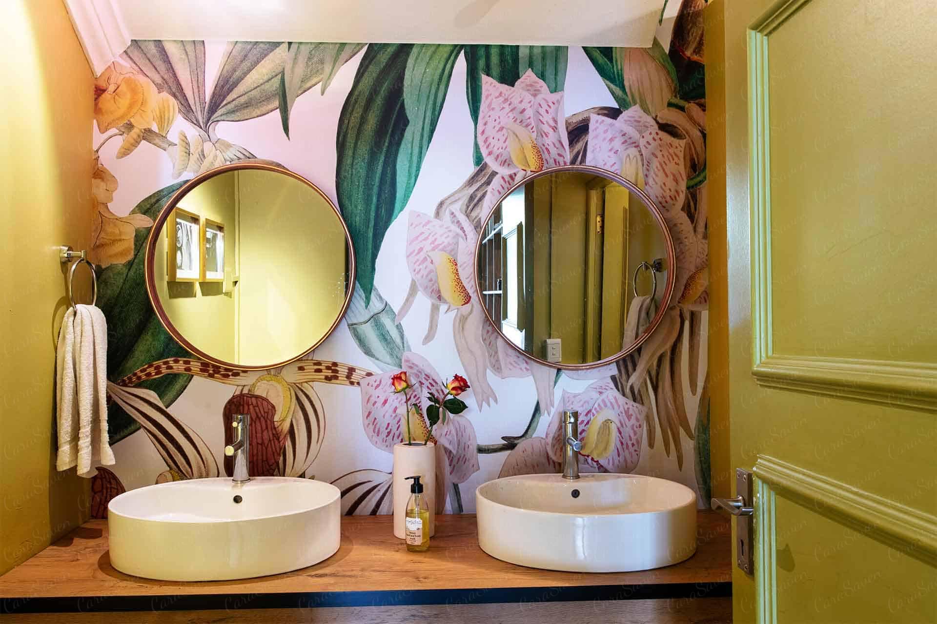 The wallpaper in the tropical-themed bathroom showcases large, colorful floral patterns featuring vibrant shades of green, pink, orange, and yellow. The detailed design includes lush leaves and exotic flowers that evoke a lively and refreshing ambience. Perfect for search terms like "large colorful floral wallpaper," "vibrant tropical wall decor," or "exotic leaf flower print.