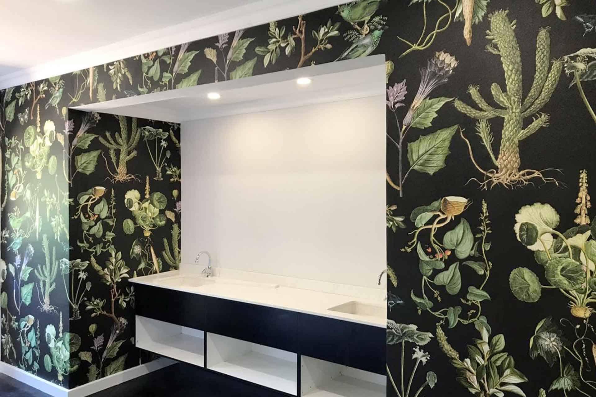 A modern room with botanical-themed wallpaper, ideal for a digital portfolio backdrop. The wallpaper showcases detailed plant illustrations: ferns, monstera leaves, palm fronds, and delicate flowers. The color palette includes rich greens, earthy tones, soft whites, and subtle yellows on a dark background.