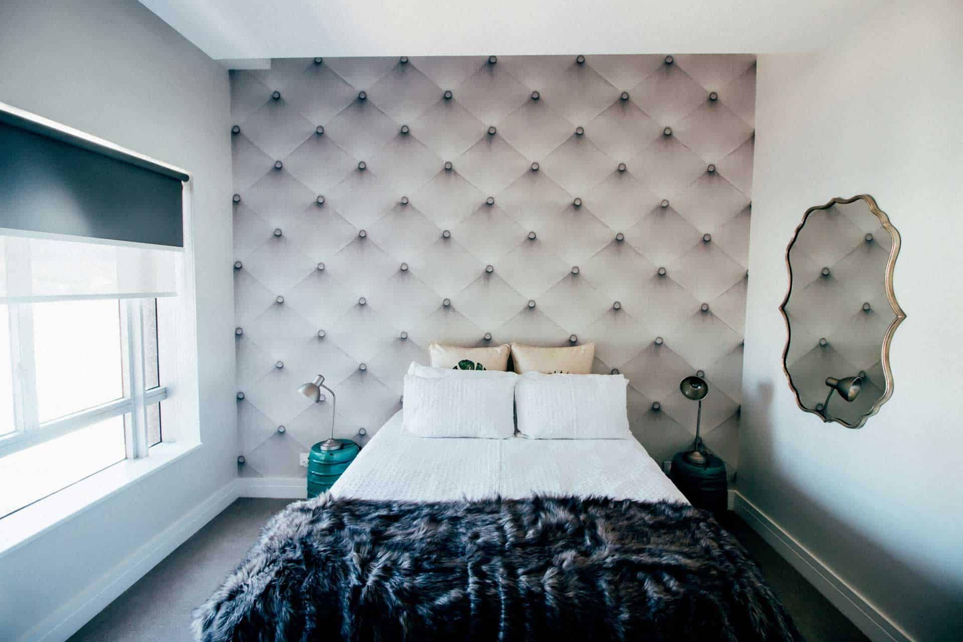 The modern bedroom features an elegant tufted accent wall behind the bed. The wallpaper boasts a plush, textured pattern with soft, neutral tones of beige and white. The tufting creates a luxurious, diamond-shaped design, adding depth and sophistication to the overall aesthetic.