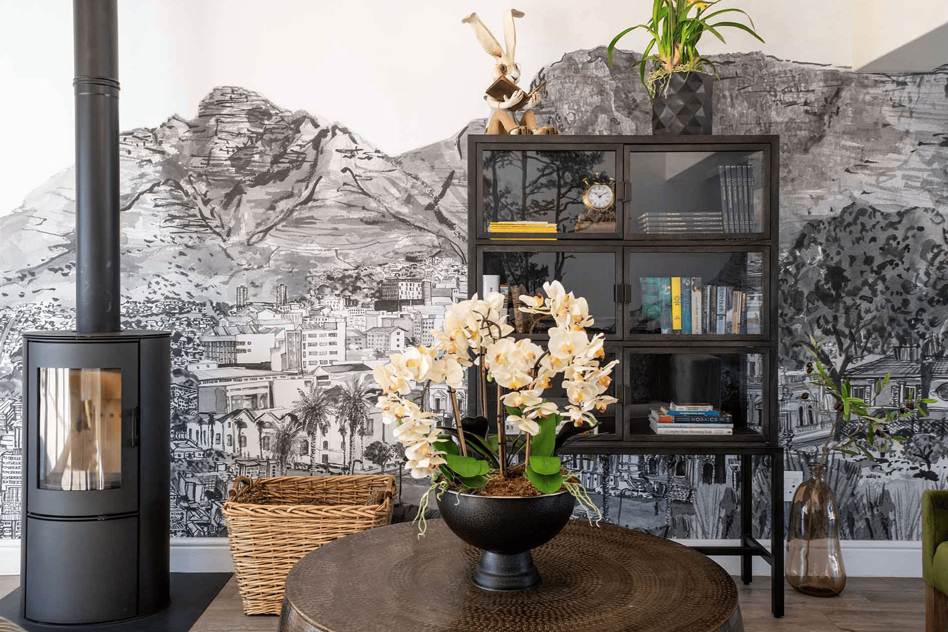 The wallpaper in the living room features a textured black-and-white cityscape mural. It includes elements like tall buildings, sharp architectural lines, and an urban skyline with varying shades of black, white, and gray. This mural gives an office wallpaper feel to the space with its detailed and monochromatic design.