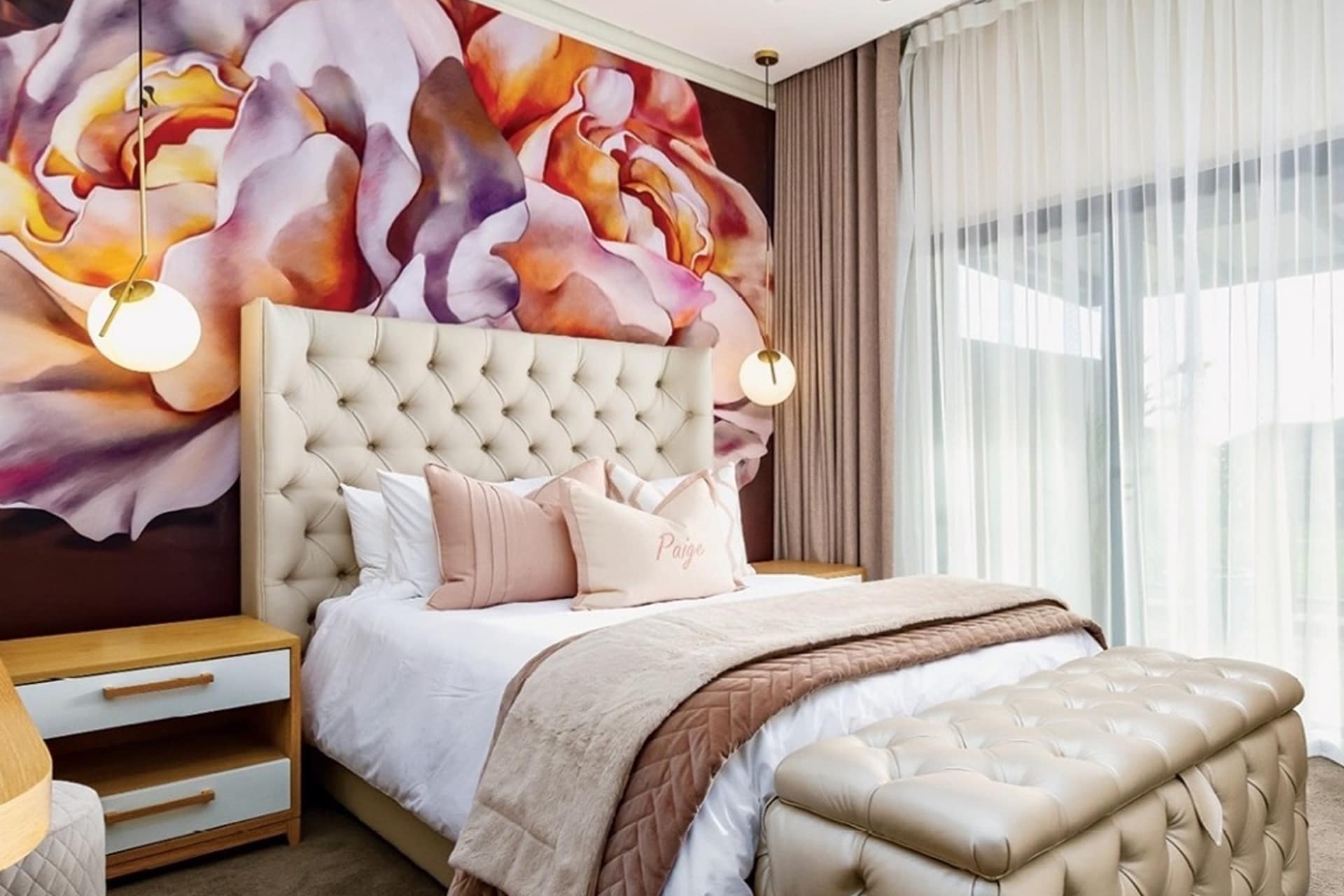 Large floral wallpaper mural with vivid, oversized flowers in shades of blush pink, soft beige petals, and deep green leaves. The background is a subtle off-white or light cream tone, enhancing the delicate yet bold floral patterns.