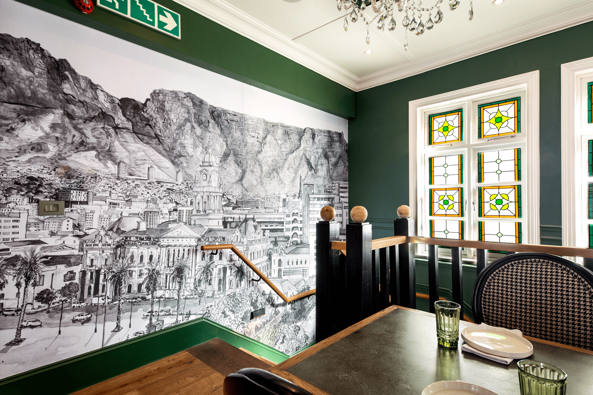 Dark green walls adorned with a large black-and-white mural of a mountainous cityscape. This striking wallpaper features intricate details of mountains and a city, offering high contrast between the dark, rich green and the monochrome mural. Ideal for making a bold statement in any room.