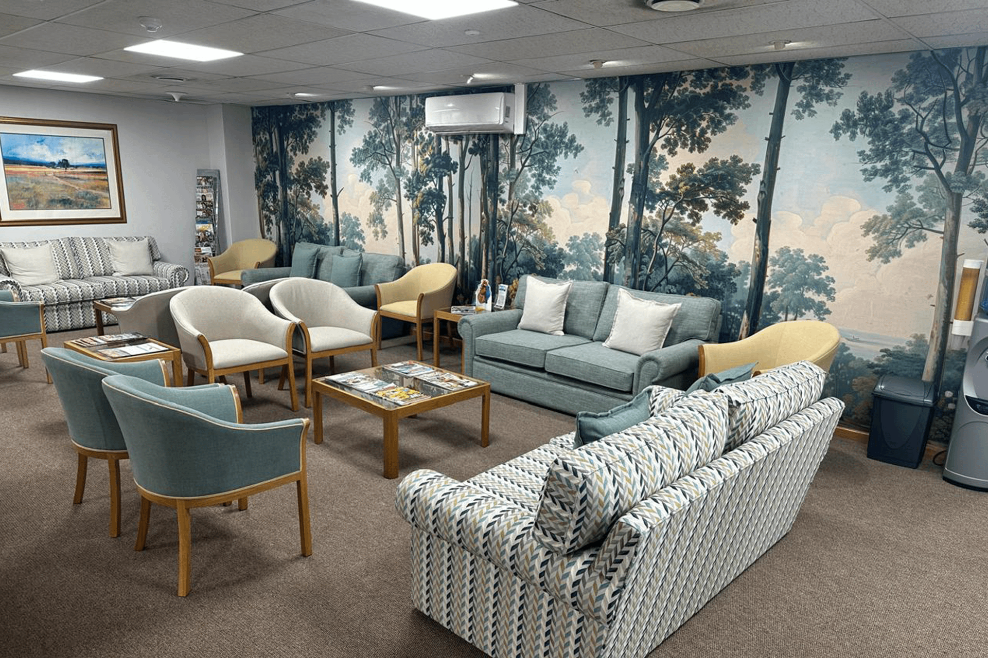 The wallpaper in the cozy waiting room is a large mural depicting trees. The mural features earthy and natural tones, primarily greens and browns, with hints of other natural colors such as muted yellows and subtle grays. These elements create a tranquil and inviting atmosphere that provides a calming backdrop to the space.