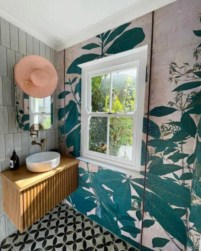 A green leafy mural envelops the wall, evoking the essence of a lush forest. Various shades of green are prominent, interspersed with hints of lighter hues resembling sunlight filtering through foliage. Delicate vines and broad leaves create an immersive natural backdrop, making it the central element in this botanical-themed decor.