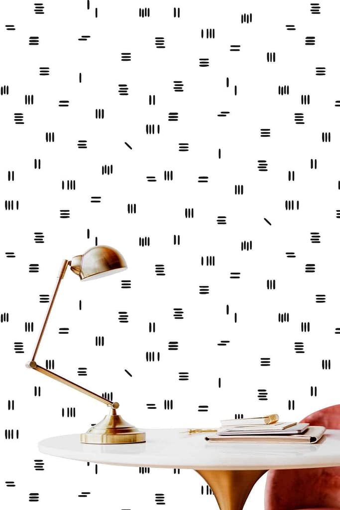 A modern workspace showcases a white desk with a brass lamp, paired with a pink chair and a stack of books. The backdrop is adorned with minimalist wallpaper featuring small black vertical and horizontal lines on a white background, evoking an elegant yet understated style akin to Simon's precise aesthetic. Touches of brass and pink complete the serene palette.