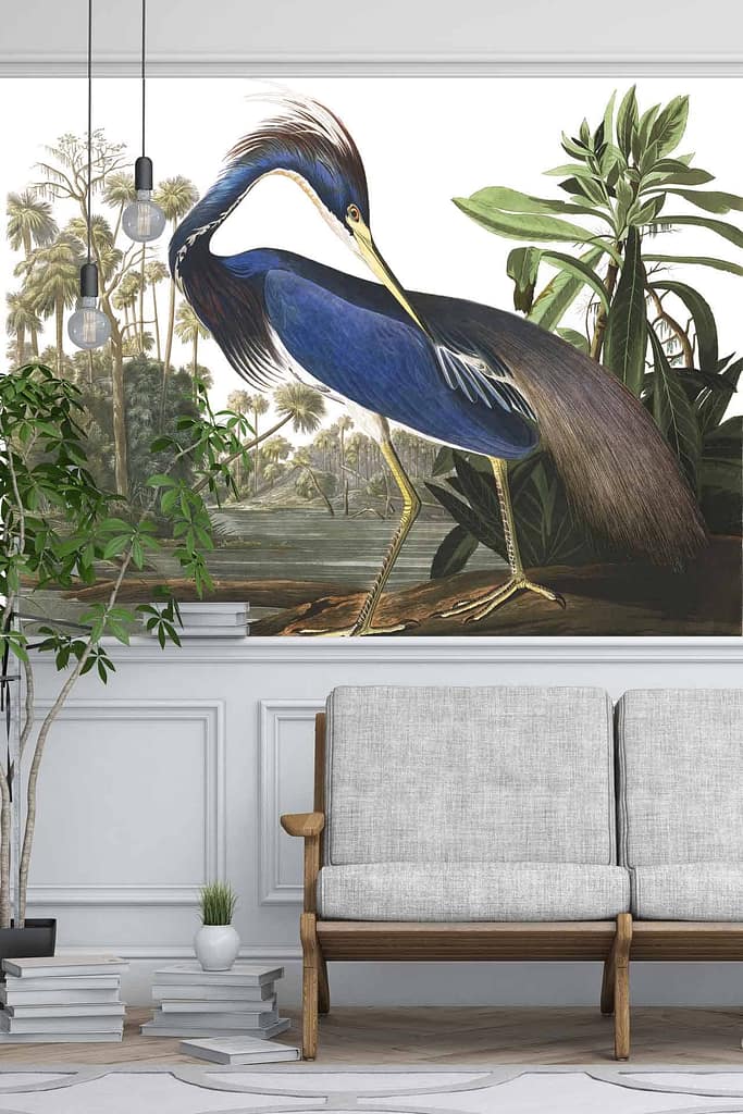 The wallpaper in the modern living room showcases a detailed mural of a blue heron, accentuated by an exotic Louisiana backdrop featuring lush trees and plants. The primary colors are shades of blue for the heron and various greens for the vegetation, with additional earthy tones in the background elements.