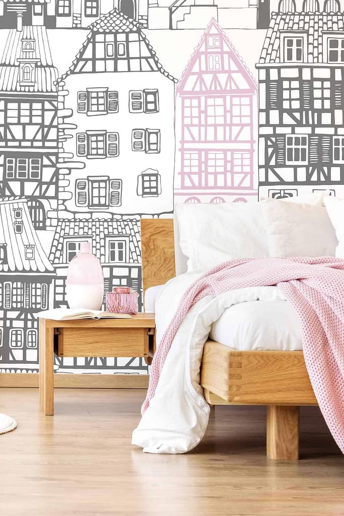 The wallpaper features a black and white mural of traditional European houses with one house highlighted in pink. The design captures the charm of playing house, combining monochromatic tones with a touch of pink for visual interest.