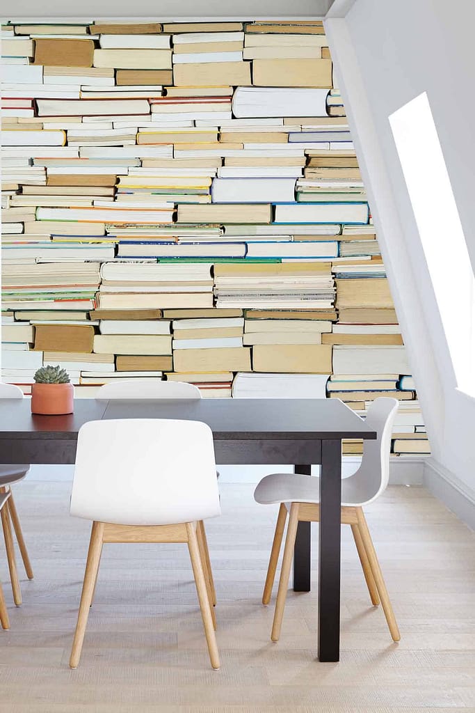 The wallpaper mural features upright and stacked books, showcasing elements such as book spines, titles, and varying heights. Dominant colors include earthy browns, rich reds, muted greens, deep blues, soft yellows, and subtle greys. The intricate details and realistic textures create a cozy literary atmosphere perfect for any reading enthusiast.