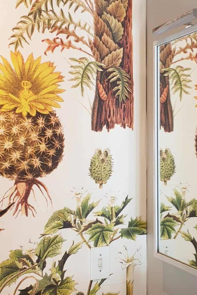 A close-up photo showcases tropical-themed wallpaper featuring vibrant botanical illustrations like sunflowers and cacti. Elements include lush green leaves, various plant textures, and bright yellow sunflower accents. The color palette boasts rich greens, sunny yellows, earthy tones, and hints of soft blues and pinks. Ideal for interior design inspiration with its lively and natural aesthetic.