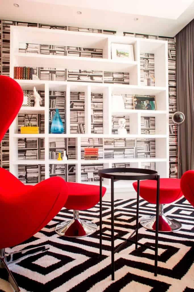 Book-themed wallpaper featuring intricate illustrations of open and closed books, shelves filled with classic literature, and vintage book spines. The color palette includes shades of beige, sepia, brown, and hints of gold or bronze detailing for an antique effect. Some sections feature subtle accents in muted reds and deep greens.