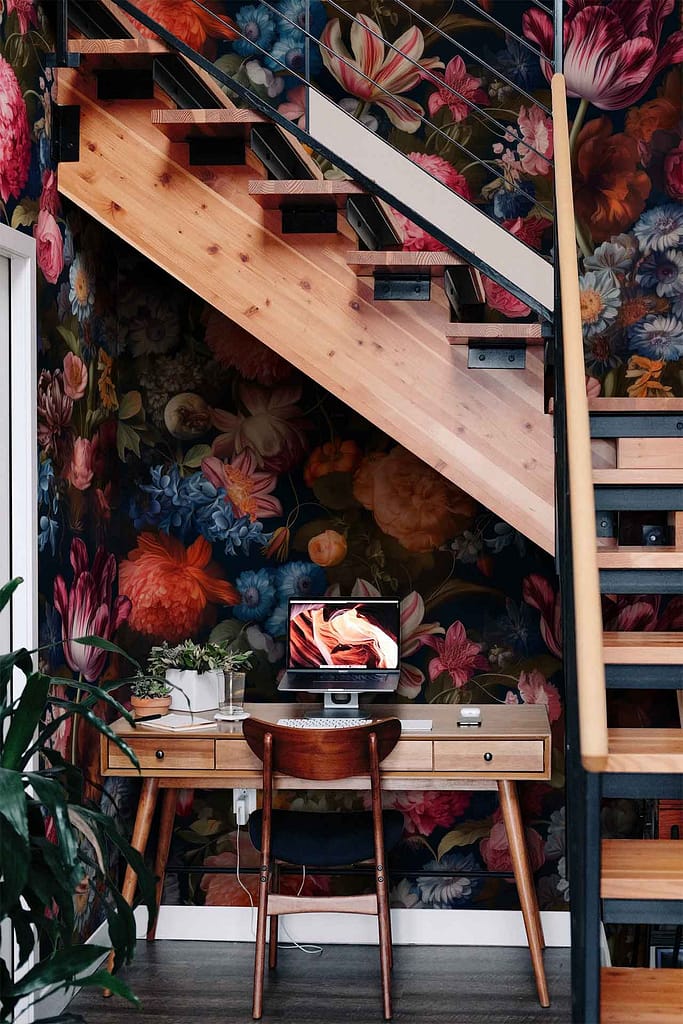 The vibrant floral wallpaper under the floating wooden staircase features elements like blooming roses, delicate peonies, and lush green leaves. The colors are rich and varied, including deep reds, soft pinks, bright yellows, and fresh greens. This lively pattern serves as a colorful backdrop for the cozy workspace setup.