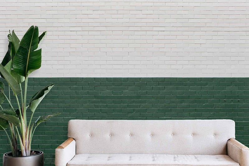 The wallpaper features a half and half design with the top section painted white and the bottom dark green, mimicking a brick wall texture. It contrasts starkly with the elegant interior elements like the white tufted sofa with wooden armrests and is complemented by lush greenery from a nearby potted plant. The clean lines and color blocks create a modern aesthetic in harmony with natural accents.