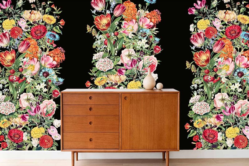 Idas Garden wallpaper features a bold, vibrant floral pattern on a black background. The design includes an array of colorful flowers in shades of red, pink, yellow, and green. Influences from nature are evident through intricate details like leaves and stems, contrasting vividly against the dark backdrop.