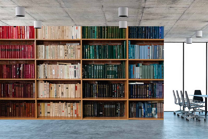 The wallpaper features a library room with a large wall-to-wall bookshelf filled with neatly arranged books. The books are sorted by color: red, beige, green, blue, and black. The room includes elements like a concrete ceiling and white cylindrical light fixtures. A sleek conference table with chairs is also visible in the design.